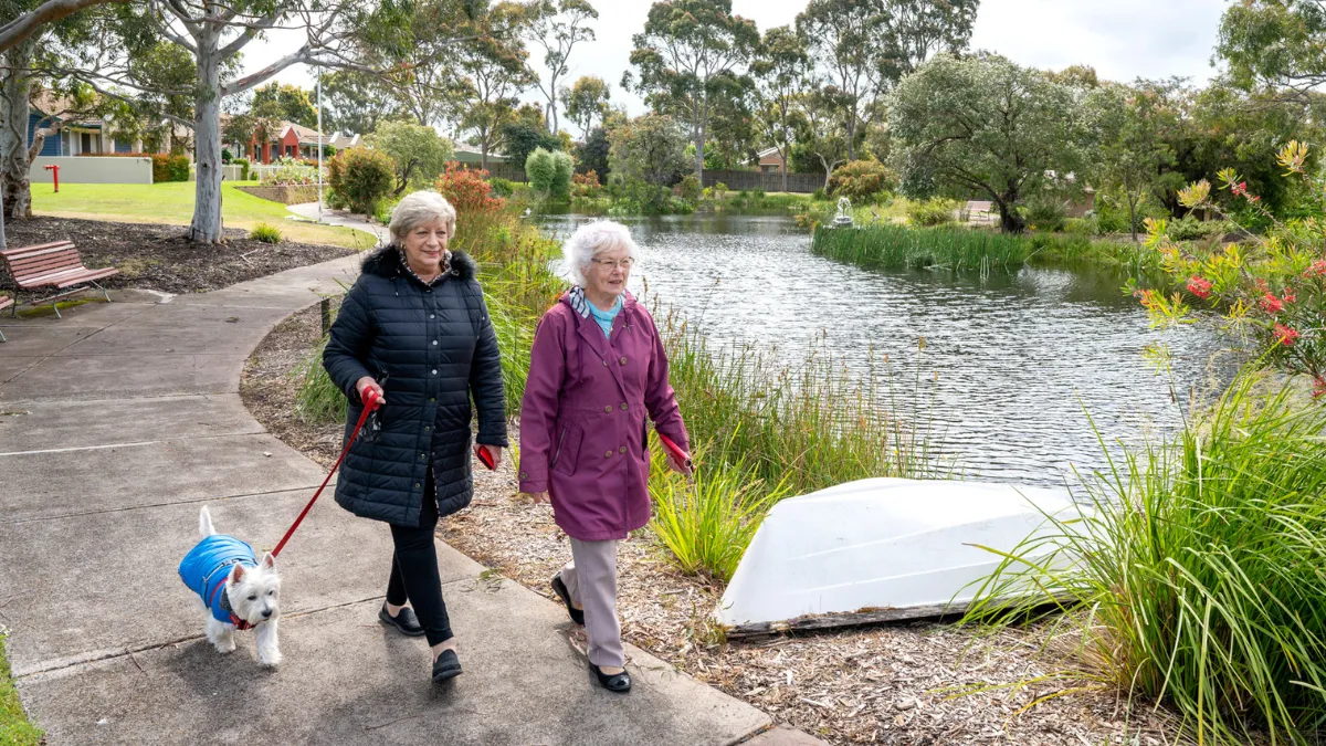 Two Female Residents Walking With A Dog Around Lake At The Mornington Retirement Village