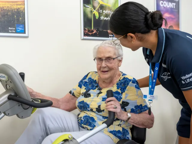 Female Resident Using An Exercise Machine With Trainer Assistance At Bucklands Residential Care