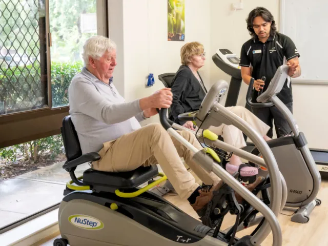 Male and Female Resident On Exercise Machines With Assistance From Male Allied Health Staff Member At Fullarton Residential Care