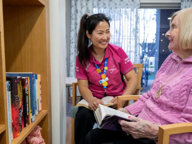 Female Resident and Female Lifestyle Coordinator Reading A Book Together In Library At Oaklands Park Lodge Residential Care