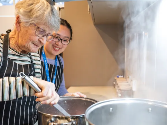 An older woman attending Southern Cross Care’s Dementia Cottage Respite services cooks on a stove top with help from a respite assistant