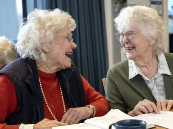 Two ladies laughing over a cup of tea and a book in aged care Adelaide, SA.
