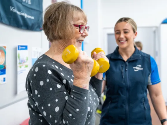 Female Client Lifting Dumbbells With Exercise Physiologist Supporting