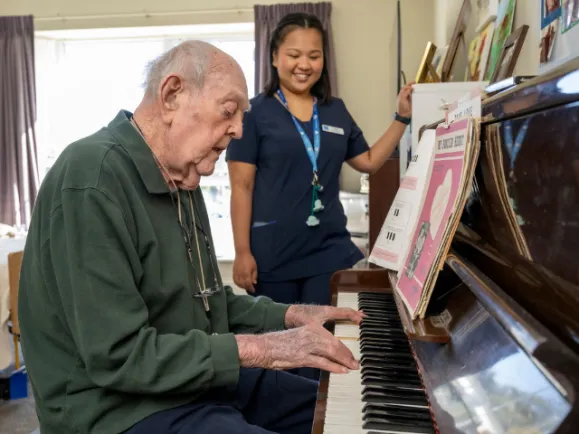 Male Resident Playing Piano and Female Staff Member Watching At The West Beach Residential Care