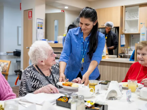 Personal Care Worker Serving Meal To Female Resident At Oaklands Park Lodge Residential Care