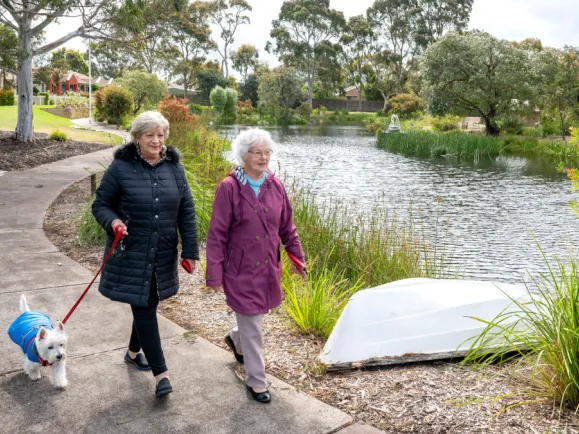 Two Female Residents Walking With A Dog Around Lake At The Mornington Retirement Village