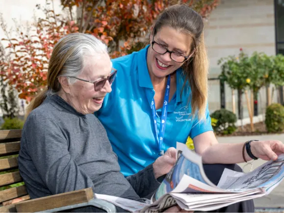 Female Volunteer Reading Newsletter With Female Resident At Southern Cross Care Residential Care Home